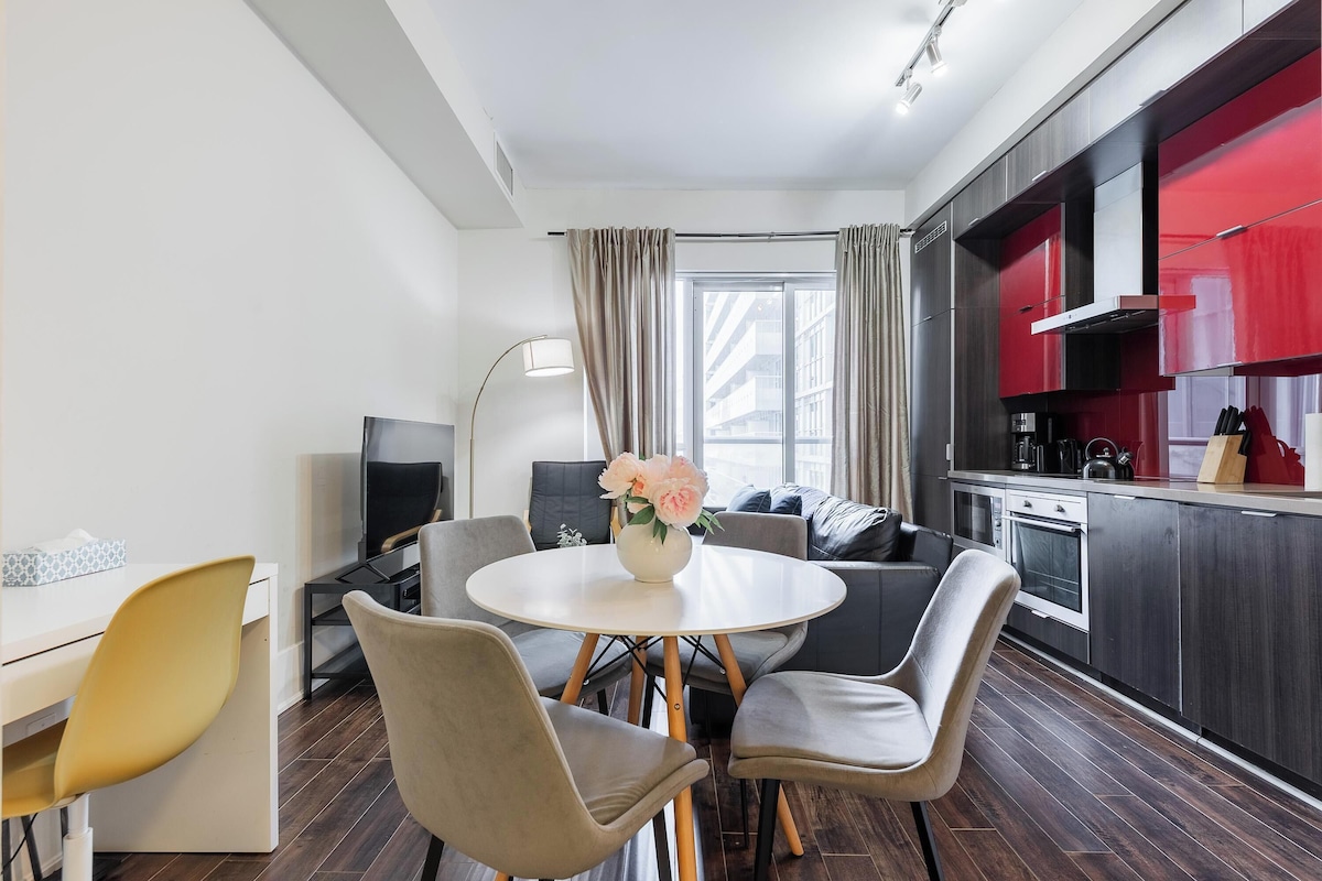 A modern kitchen with dark cabinets and a red backsplash, a dining area with a round table and chairs, and a large window with curtains.