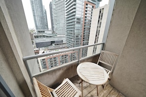 Varanda com mesa e cadeira de madeira, grade de metal e vista para edifícios altos.