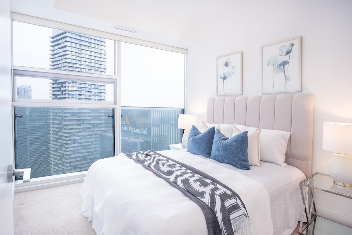 Un dormitorio moderno con un ventanal grande, una cama con colcha blanca y gris estampada, y dos cuadros enmarcados en la pared.
