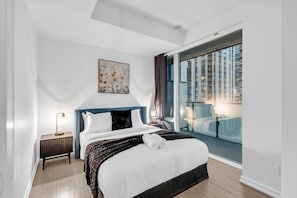 Quarto de hotel moderno com uma cama grande, uma mesa de cabeceira, uma luminária e uma janela com vista para edifícios.