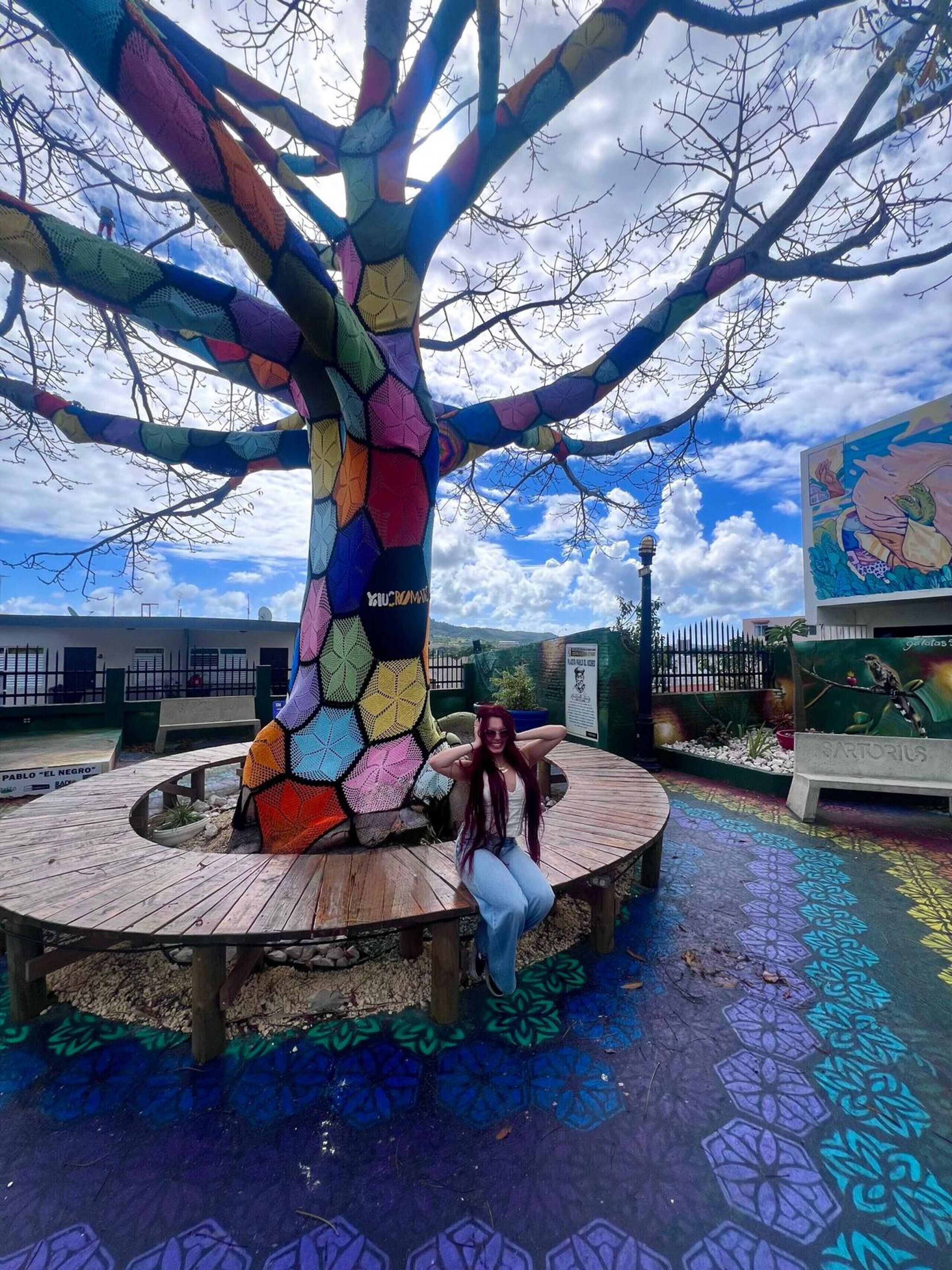 A tree sculpture with a colorful geometric pattern, a circular wooden platform, and a mural in the background.