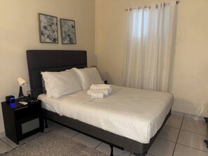 A neatly made bed with white bedding, a dark headboard, and a nightstand with a lamp and clock.