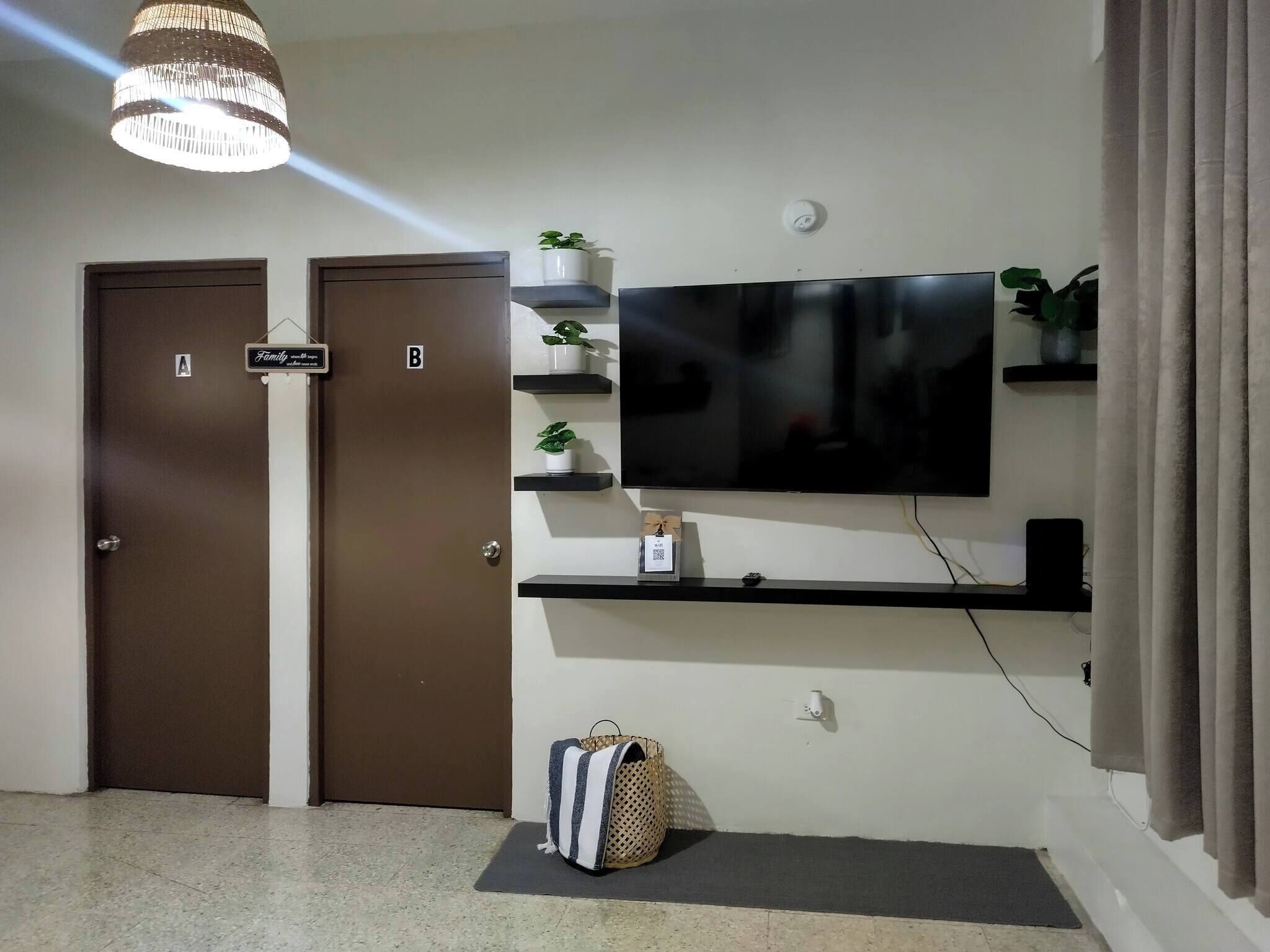 Ein modernes Wohnzimmer mit einem Flachbildfernseher, zwei Türen mit der Beschriftung 'A' und 'B', einer kleinen Pflanze auf einem Regal und einer gewebten Tasche auf dem Boden.