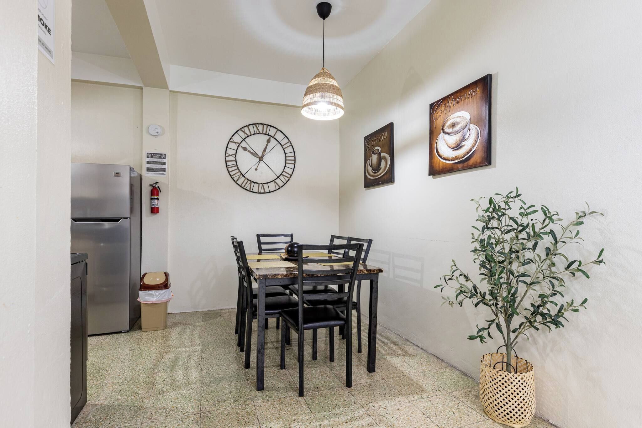 Un coin repas avec une table ronde, des chaises, une horloge, des œuvres d’art murales, un luminaire suspendu, une plante en pot et un réfrigérateur.