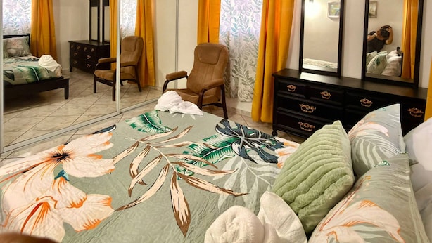 A bedroom with a bed, two chairs, a dresser, a mirror, and a window with curtains.
