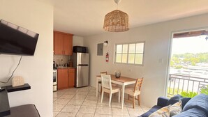 A modern kitchen with a refrigerator, microwave, and wooden cabinets. There is a dining area with a table and chairs, and a sofa in the living room. A television is mounted on the wall, and a window provides a view of the outside.
