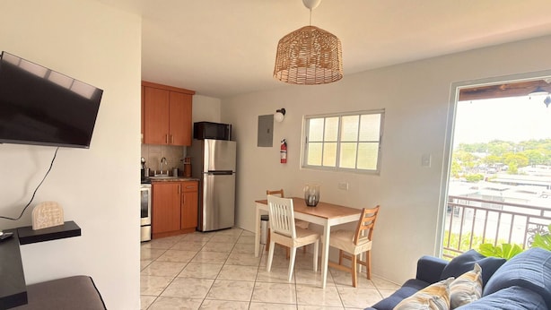 Eine moderne Küche mit Kühlschrank, Mikrowelle und Holzschränken. Es gibt eine Essecke mit Tisch und Stühlen, sowie ein Sofa im Wohnzimmer. Ein Fernseher ist an der Wand montiert und ein Fenster bietet einen Blick nach außen.