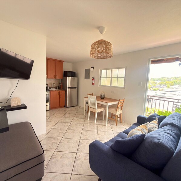 A modern kitchen with a refrigerator, microwave, and wooden cabinets. There is a dining area with a table and chairs, and a sofa in the living room. A television is mounted on the wall, and a window provides a view of the outside.