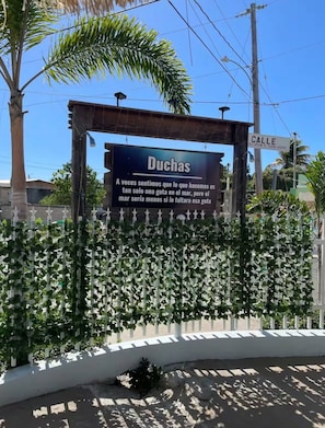 Un letrero con el nombre 'Duchas' y un mensaje en español, una cerca blanca con plantas trepadoras, y una señal de calle que dice 'Calle'.