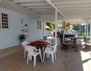 Eine überdachte Terrasse mit einem Holztisch, weißen Plastikstühlen, einer Tür und einem Blumentopf.