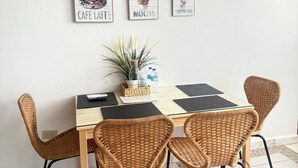 A dining area with a table, wicker chairs, a potted plant, and wall art depicting coffee drinks. - Brisas Apt | Comfort & WiFi with A/C (Yauco)