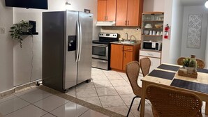 A compact kitchen with a stainless steel refrigerator, wooden cabinets, a microwave, and a dining area with wicker chairs. - Brisas Apt | Comfort & WiFi with A/C (Yauco)