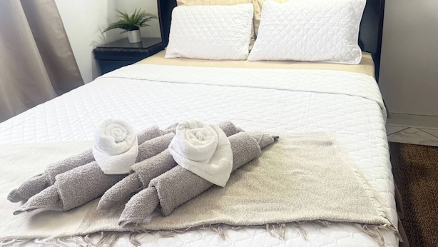 A neatly made bed with white bedding and two pillows, a dark headboard, and a small potted plant on a bedside table. - Brisas Apt | Comfort & WiFi with A/C (Yauco)