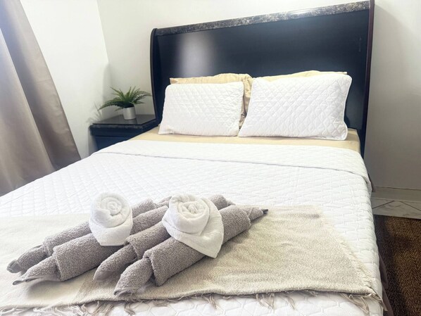 Una cama bien hecha con ropa blanca y dos almohadas, un cabecero oscuro y una pequeña planta en maceta sobre una mesita de noche.