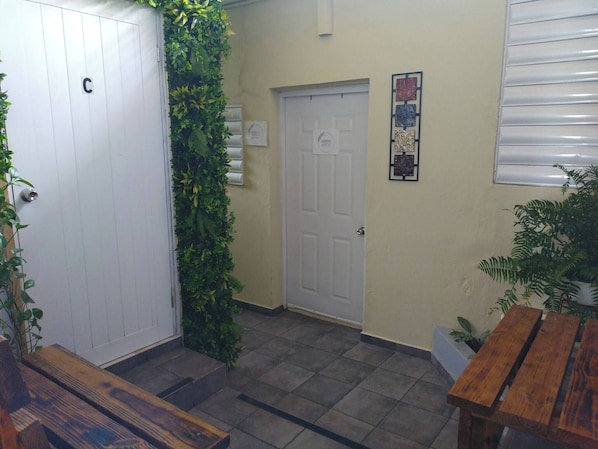 Un pasillo con una puerta blanca, un muro verde con plantas, un banco de madera y un suelo de baldosas.