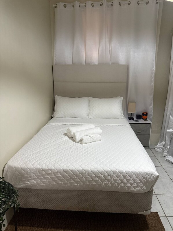 A neatly made bed with a quilted mattress, a bedside table with a lamp, and a curtain.