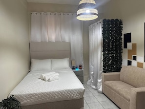 Un dormitorio ordenado con cama, mesita de noche con lámpara y una silla.