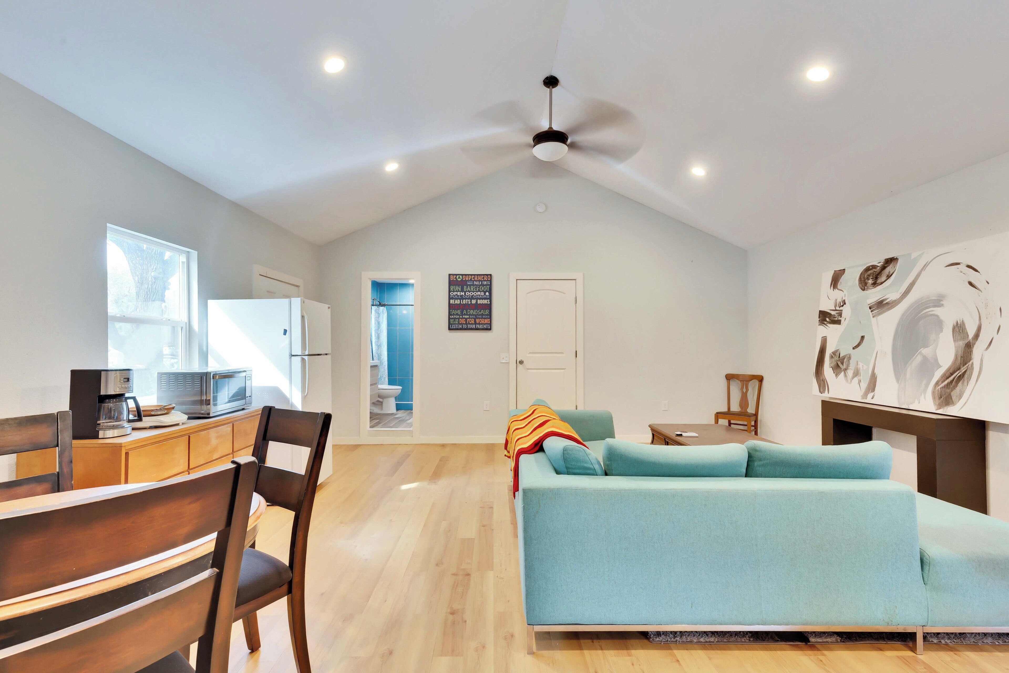 A modern living room with a sofa, dining table, chairs, a ceiling fan, and a kitchen area with appliances.