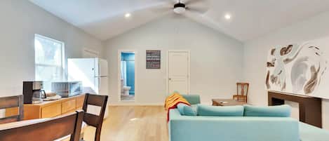A modern living room with a sofa, dining table, chairs, a ceiling fan, and a kitchen area with appliances.