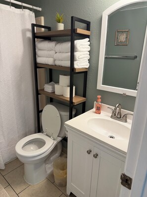 A bathroom with a toilet, a sink, a mirror, a towel rack, and a shelf with towels and toilet paper. -  1-bedroom condo in South Padre Island with views of the beach, and Laguna Madre (South Padre Island)