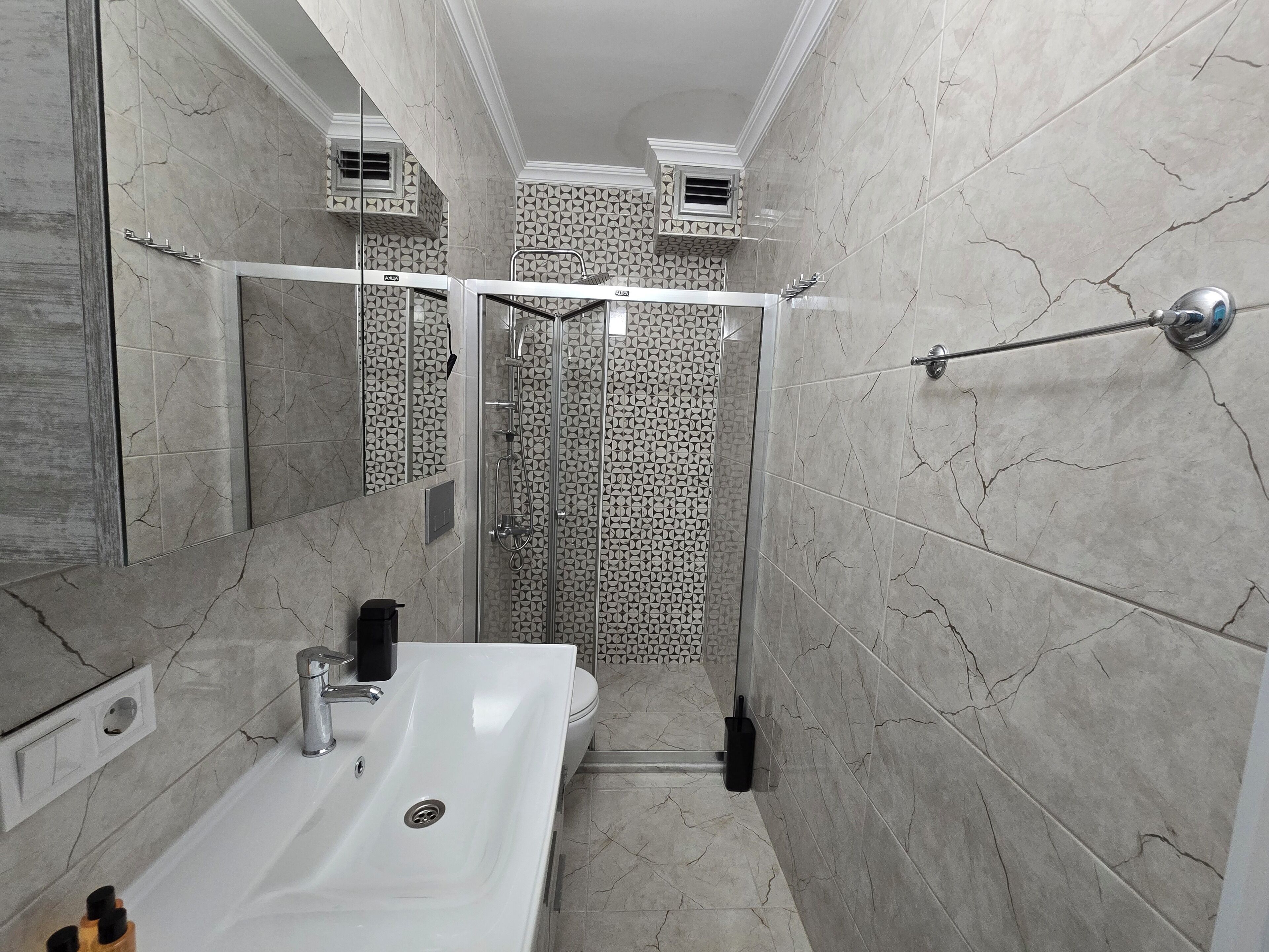 Een badkamer met een witte wastafel, een spiegel, een douche met glazen deur en betegelde muren.