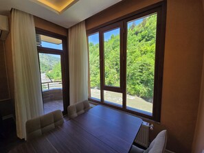 A room with a large window overlooking a river and forest, a wooden table, and a chair. - Esla Suites (Çamlıhemşin)