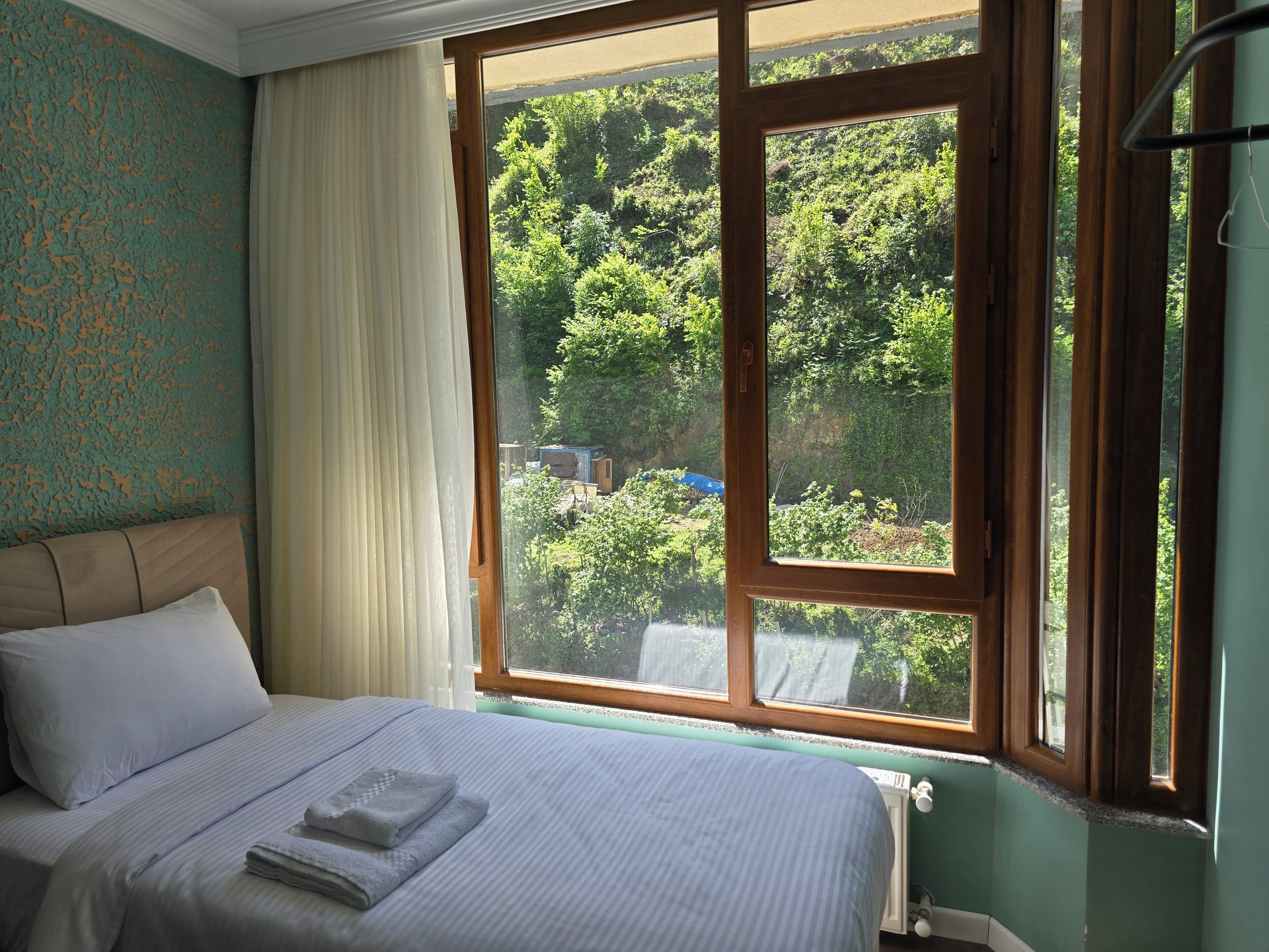 Habitación de hotel con un ventanal grande, cama con ropa blanca y vistas a un área boscosa.