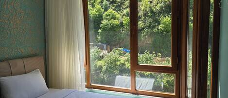 Habitación de hotel con un ventanal grande, cama con ropa blanca y vistas a un área boscosa.
