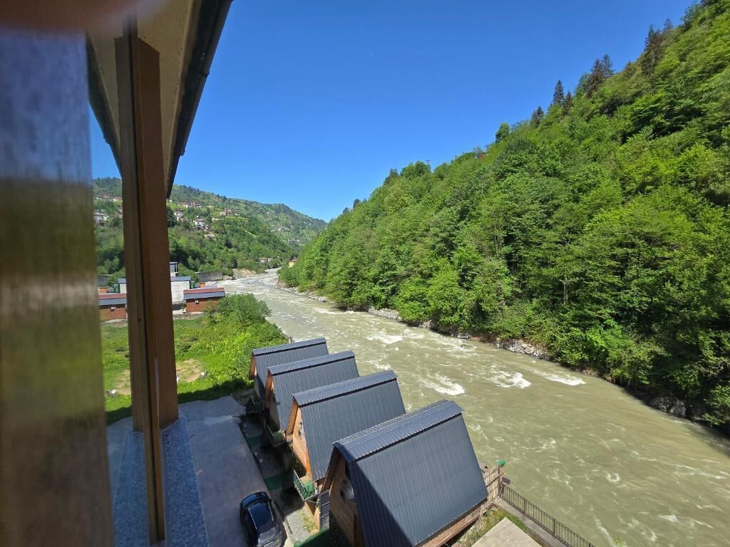 Une rivière traversée par un pont en bois, une rangée de cabanes et une colline boisée.