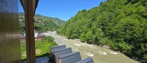 A river with a wooden bridge, a row of cabins, and a forested hillside.