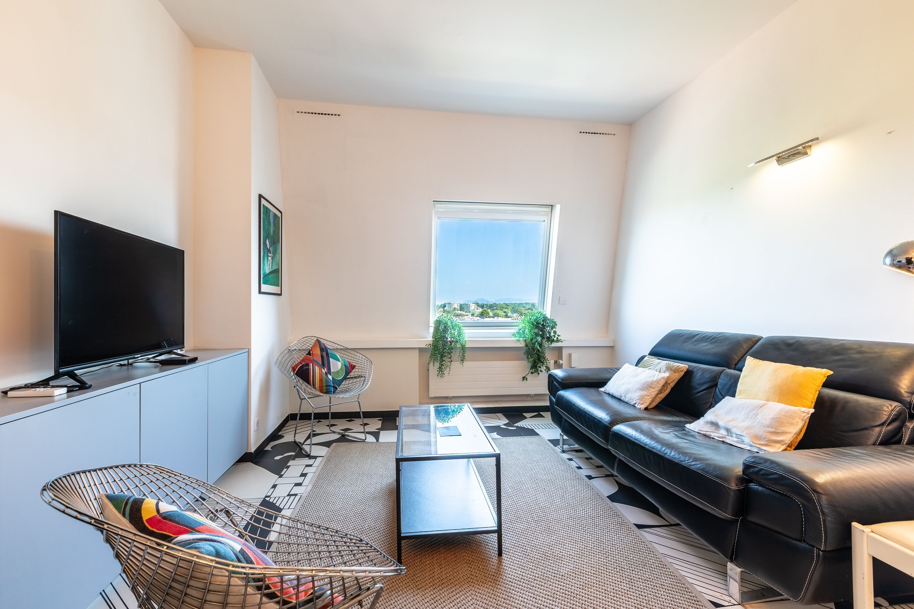 A modern living room with a flat-screen TV, a glass coffee table, a black leather sofa, and a patterned rug.