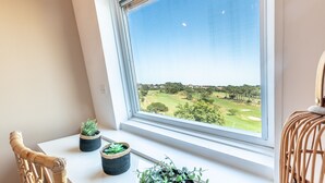 Ein Fenster mit Blick auf einen Golfplatz, ein Korbsessel und zwei Blumentöpfe auf einem Fensterbrett.