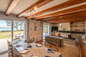 A dining area with a table set for four, wooden beams on the ceiling, a view of the outdoors, and a kitchen area with shelves. - Maison de la Falaise - Accès direct à la plage (Pénestin)