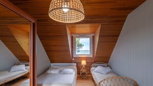 Um quarto com tetto inclinado de madeira, uma pequena janela com vista, duas camas com roupa de cama branca e uma luminária de fibra trançada.