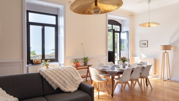 A dining area with a table set for four, a sofa with a white throw, and large windows with a view. - La chevreuse - Vue mer à moins de 300m de la plage (Lamballe)