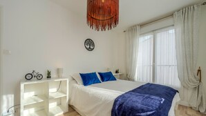 A bedroom with a bed, white nightstands, a white shelving unit, a hanging light fixture, and a window with curtains. - La Terrasse de la Rade - Terrasse avec vue mer (Brest)