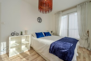 Ein Schlafzimmer mit einem Bett, weißen Nachttischen, einem weißen Regal, einer Pendelleuchte und einem Fenster mit Vorhängen.