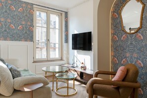 Ein Zimmer mit einer Sitzecke, einem Sessel, einem Fernseher, einem kleinen Tisch und einem Fenster mit Blick auf Gebäude.