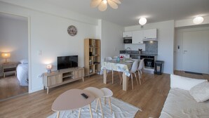Ein modernes Wohnzimmer mit Fernseher, Sitzgelegenheit, Esstisch und einer Küche im Hintergrund.