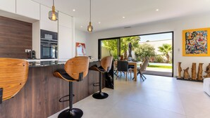 A modern kitchen with bar stools, a dining area with a view, and a living space with a large window. - Villa Camarines, new villa, pool, walk to beach (Agde)