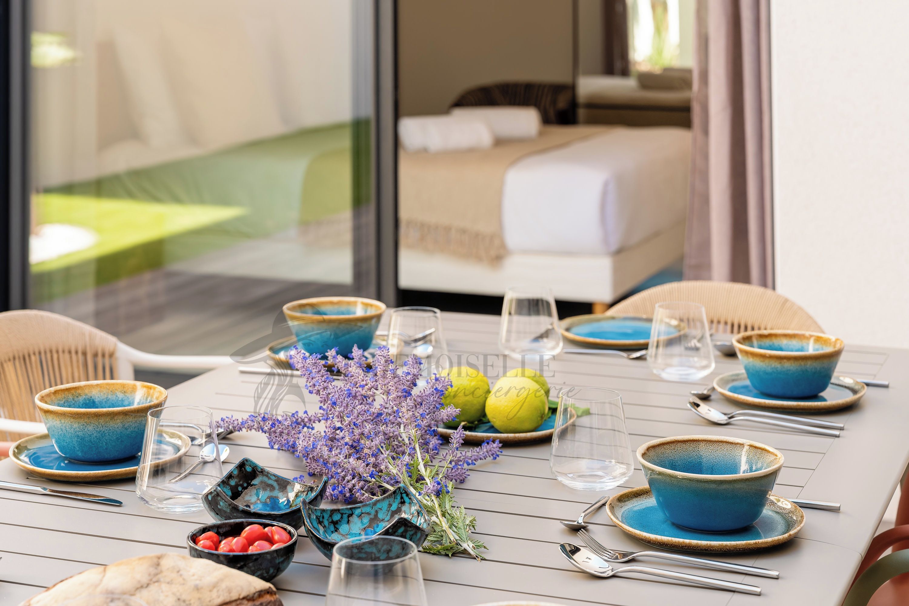 A dining table set with blue and turquoise bowls, plates, and cutlery, accompanied by a vase of purple flowers and lemons.