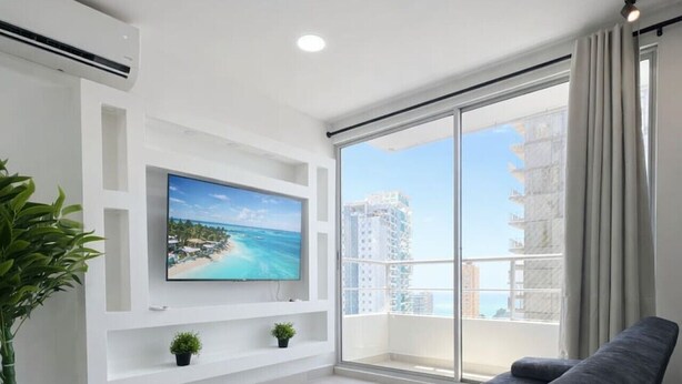 A modern living room with a large flat-screen TV, a grey sofa, a potted plant, and a balcony with a city view. - Caribbean Mist by Hilal - Wifi - Seaview - Beach (Cartagena)