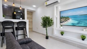 A modern living room with a sofa, a TV displaying a beach scene, a kitchen area, and a potted plant. - Caribbean Mist by Hilal - Wifi - Seaview - Beach (Cartagena)
