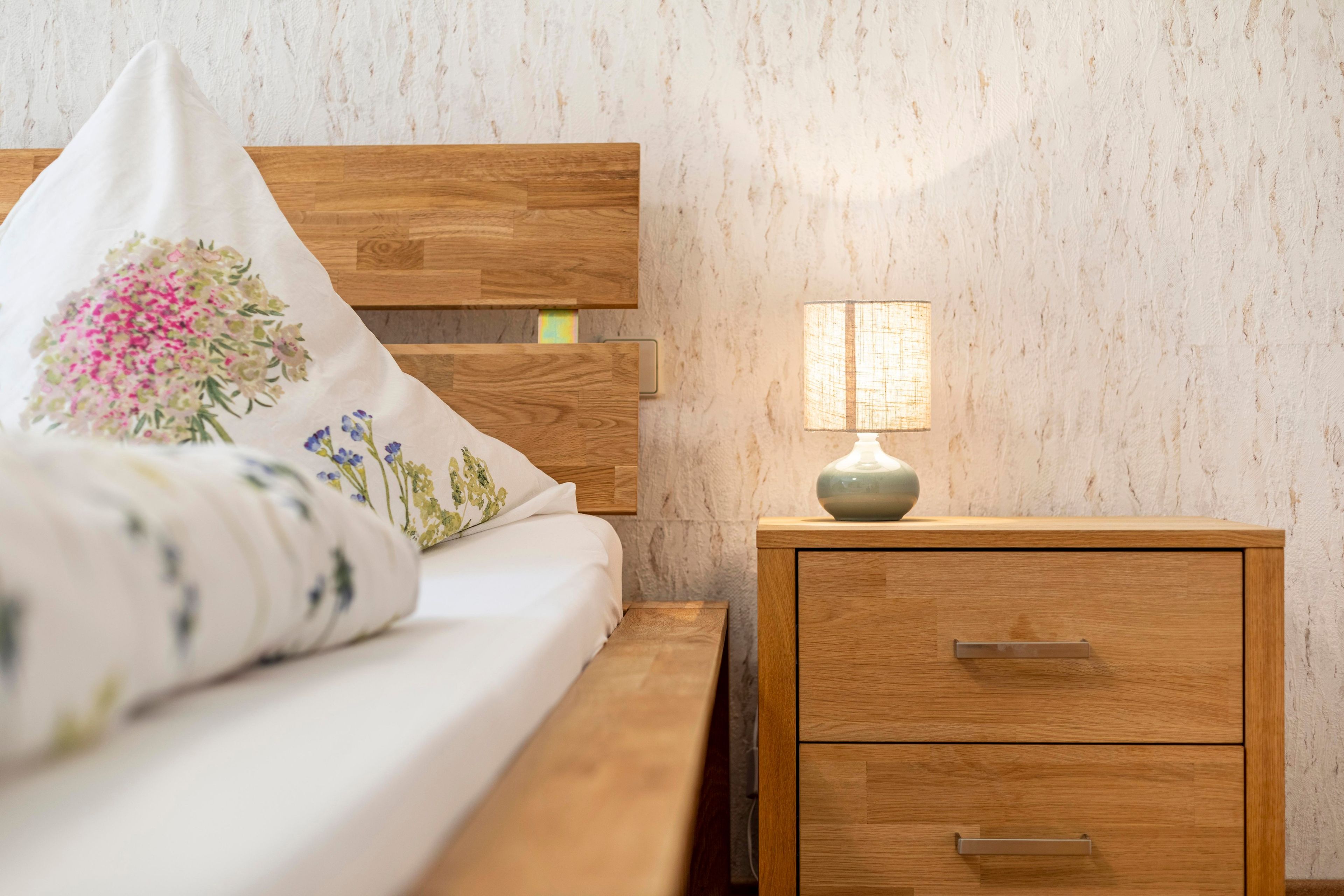 A neatly made bed with floral pillows, a bedside lamp, and a wooden nightstand with drawers.