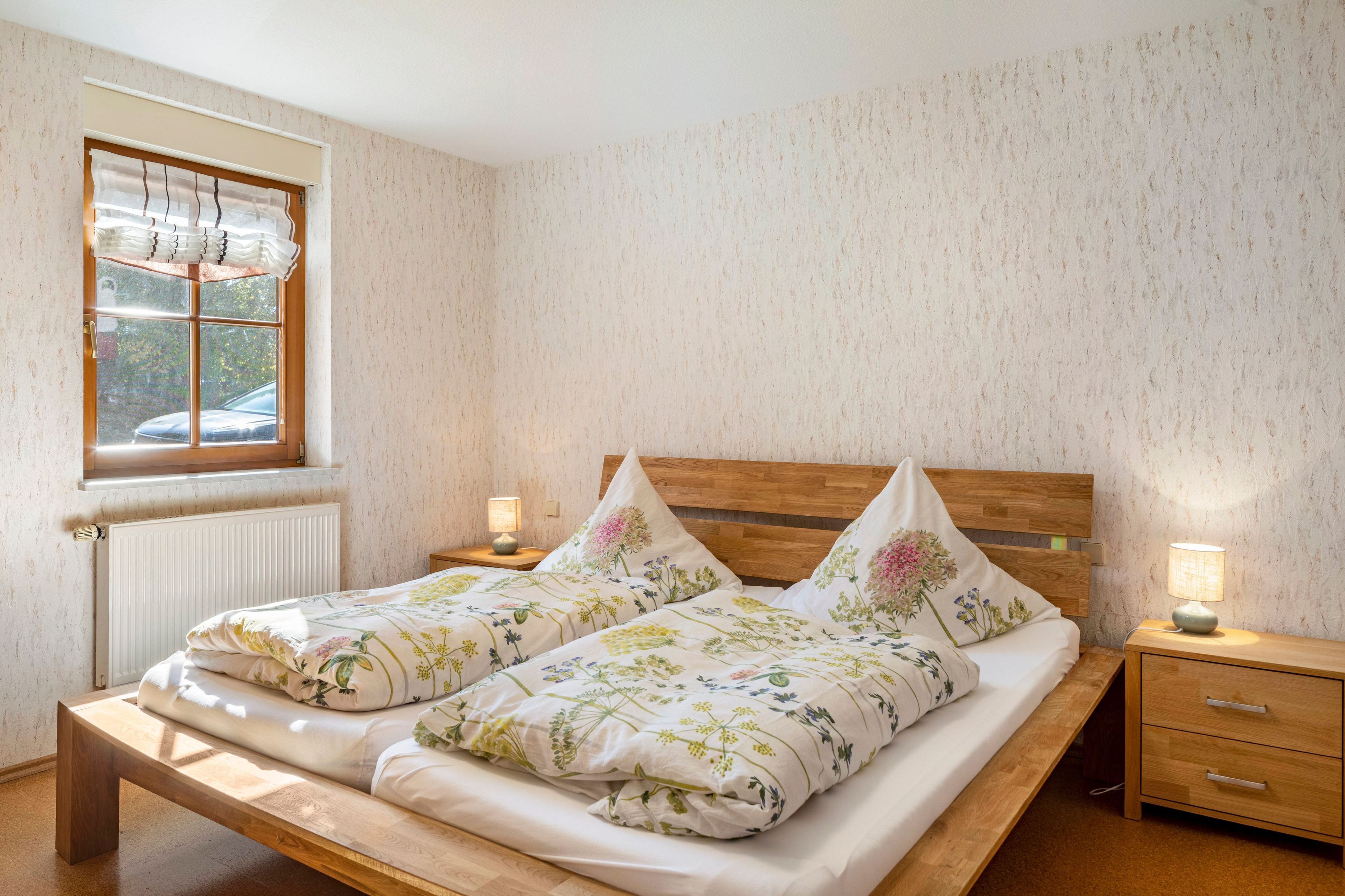 A double bed with floral bedding, a wooden headboard, bedside tables with lamps, and a window with a view of trees.