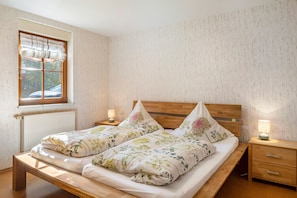 Ein Doppelbett mit floralem Bettbezug, ein Holzkopfende, Nachttische mit Lampen und ein Fenster mit Blick auf Bäume.