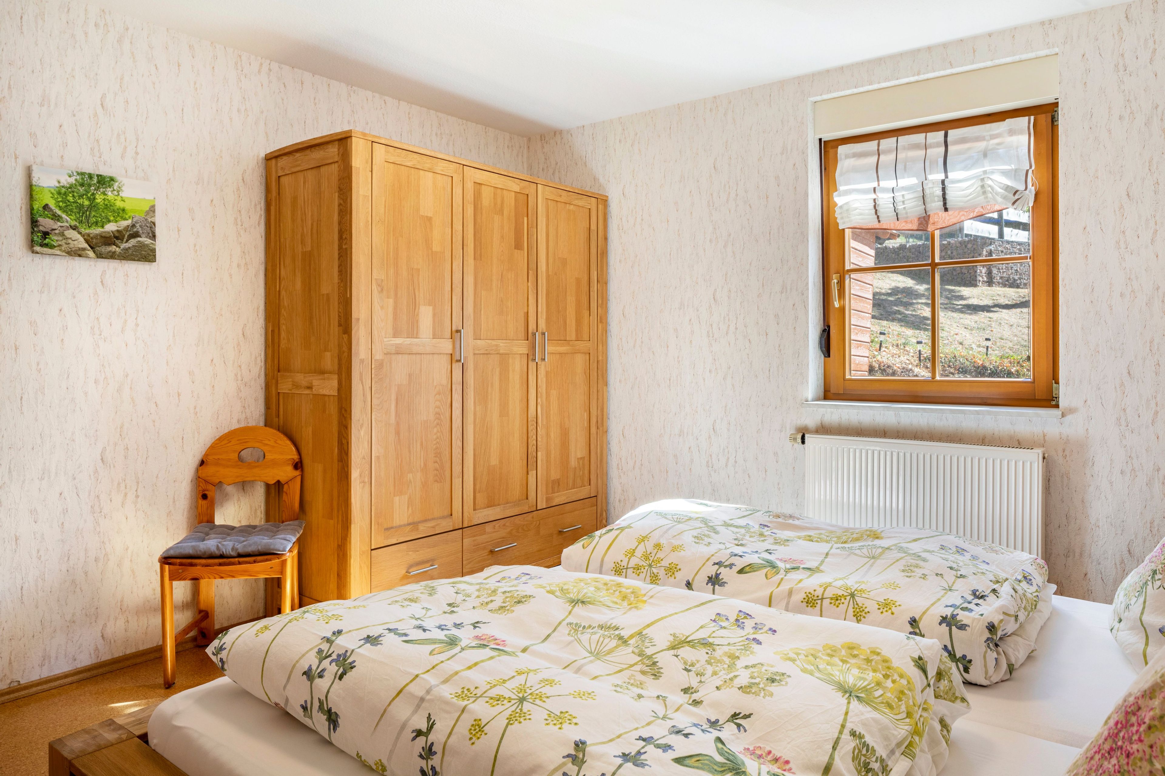 A bedroom with two beds, a wooden wardrobe, a chair, a window with curtains, and a radiator.