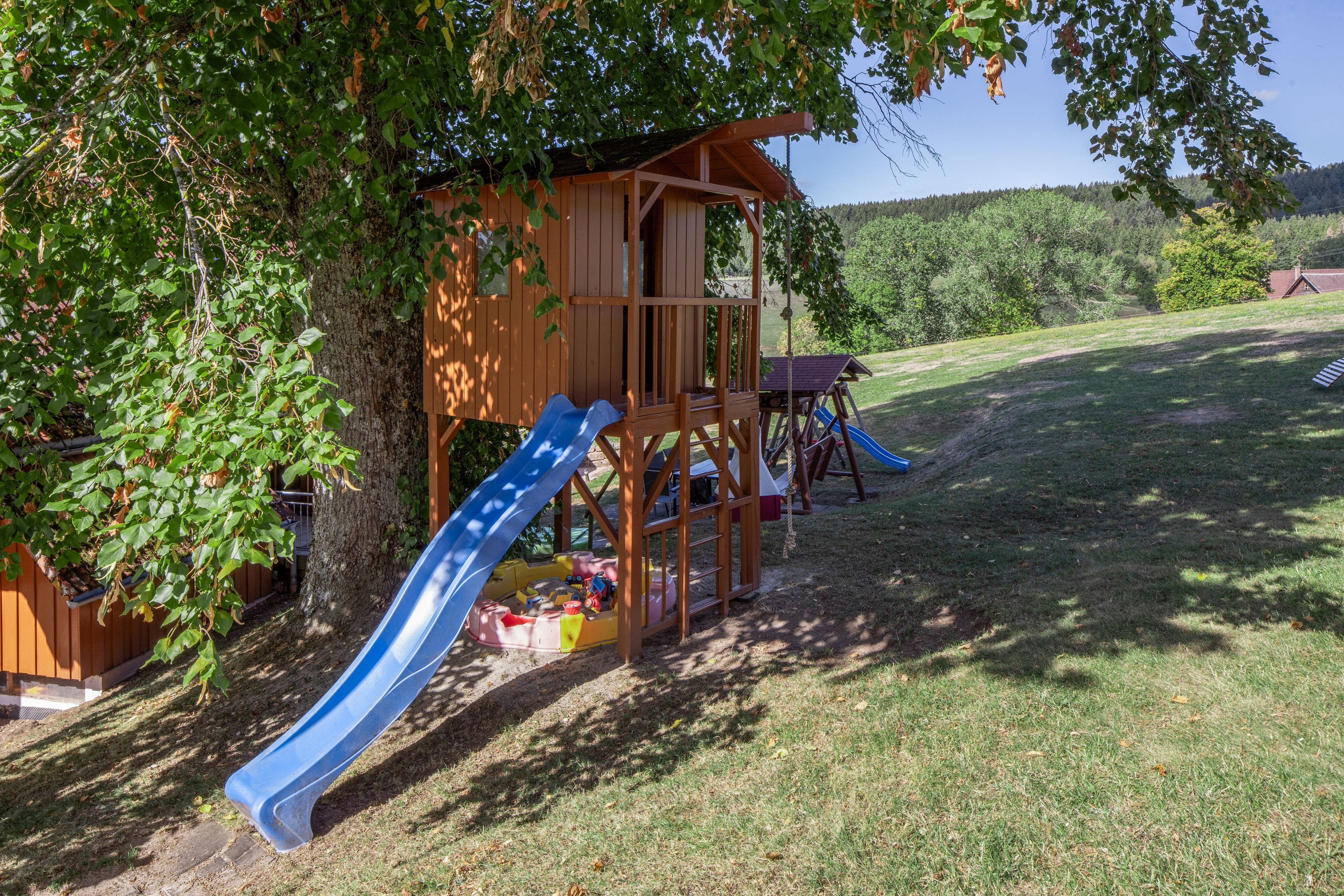 A wooden play structure with slides and climbing elements, surrounded by grass and trees.