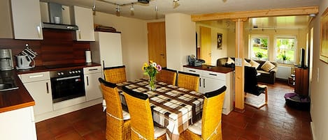 A compact kitchen with a dining area, featuring a checkered tablecloth, wicker chairs, and a central vase with flowers.
