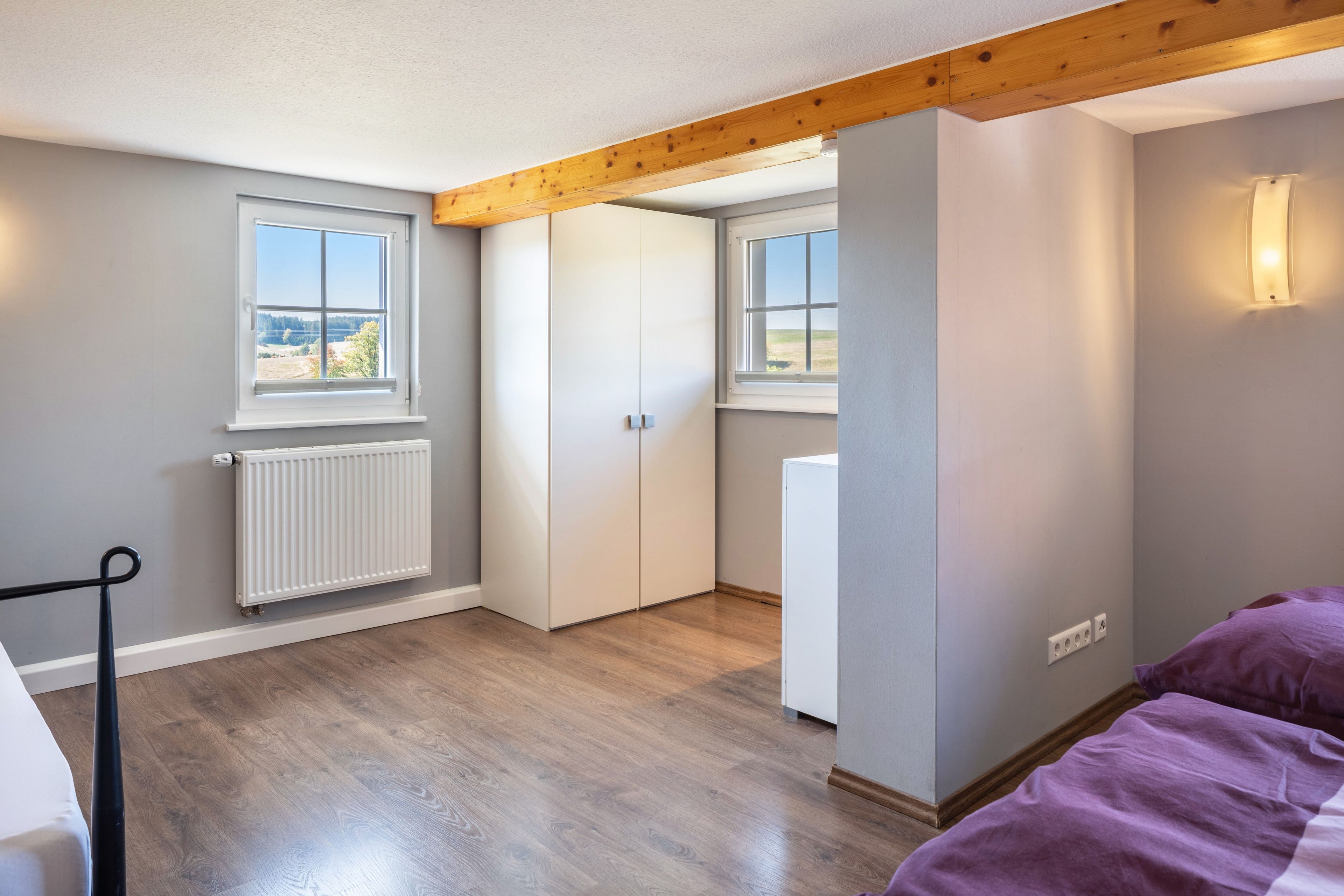 Une pièce avec un plafond en bois, un radiateur, un lit recouvert d’une couverture violette et une armoire blanche.